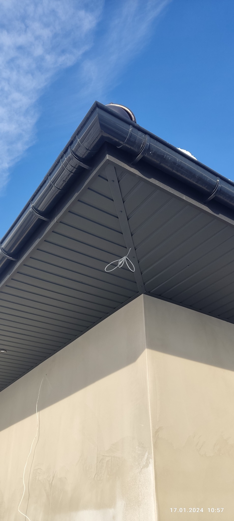 Niski kąt widzenia na narożnik dachu z czarną rynną i szarą blachą trapezową, przymocowaną do ściany budynku w słoneczny dzień. Biały sznurek zwisa z dachu.