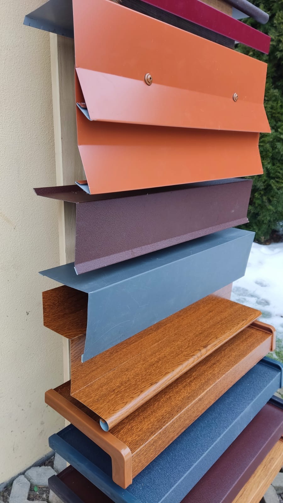 Ekspozycja różnych profili i kolorów metalowych elementów wykończeniowych dachu, w tym imitacji drewna, prezentowana na zewnątrz budynku z fragmentem zieleni w tle.