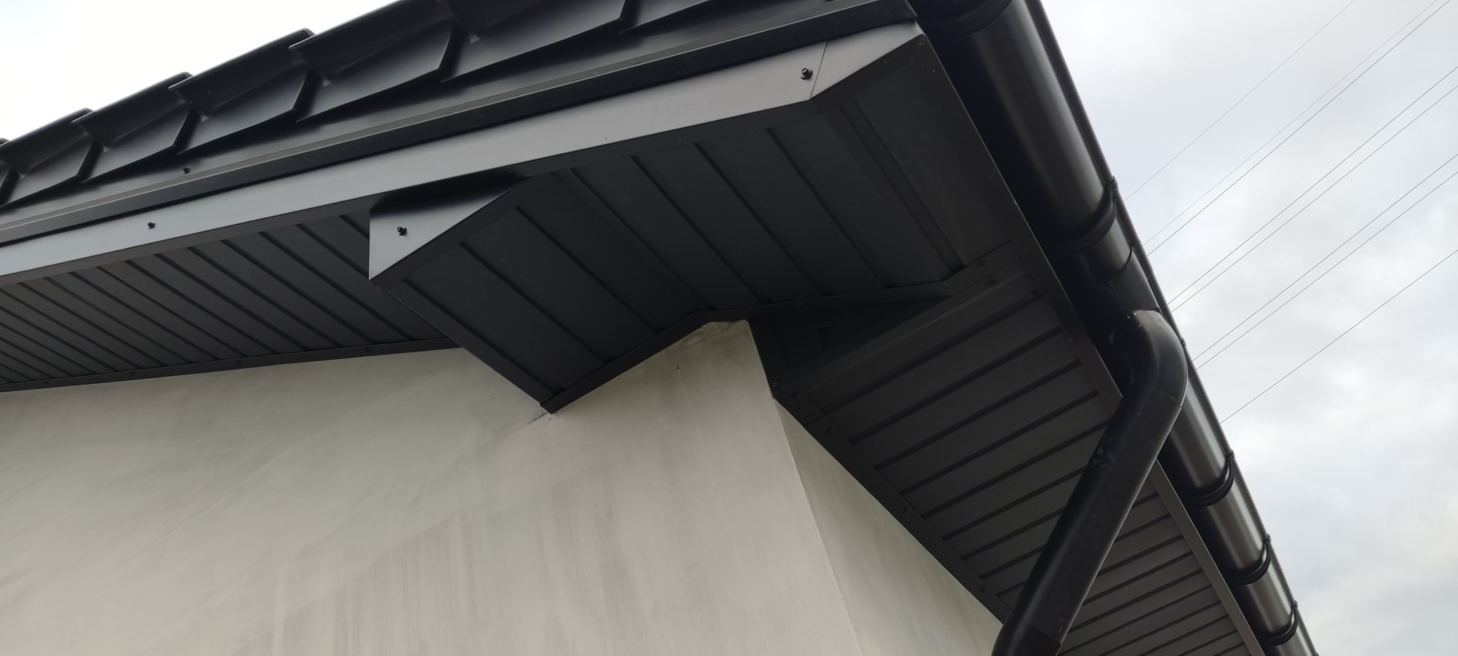 Szczegółowe ujęcie fragmentu dachu z ciemną blachą, widoczna rynna i obróbka blacharska przy ścianie budynku, na tle pochmurnego nieba.