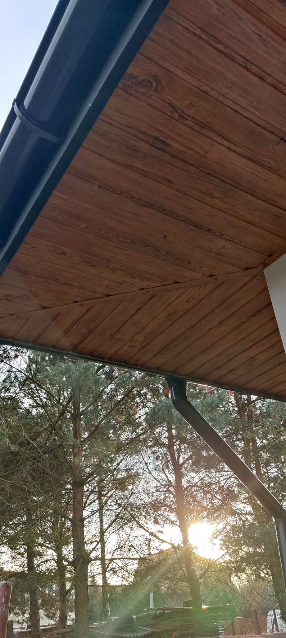 Podbitka dachowa z brązowego drewna, zamontowana rynna w kolorze grafitowym, widok z dołu na tle drzew i zachodzącego słońca.