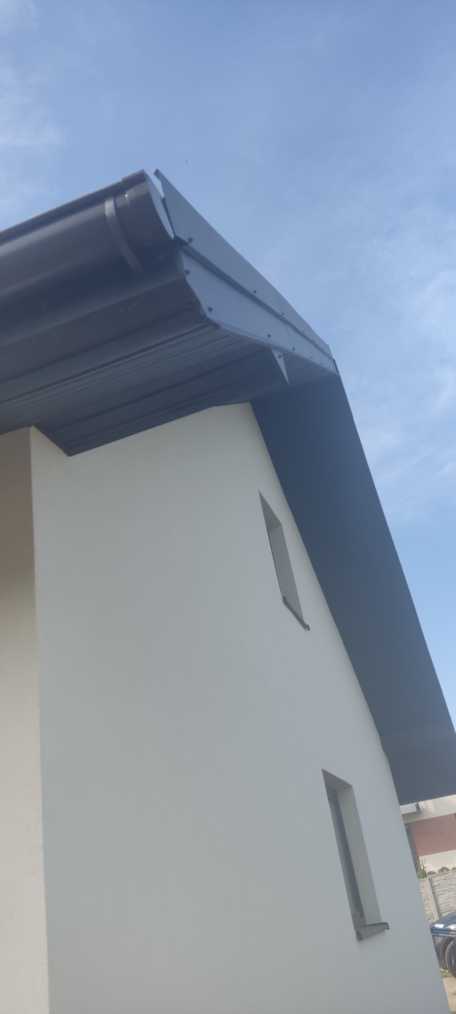 Fragment domu z nowym, grafitowym dachem z blachy, rynną i obróbkami blacharskimi, na tle błękitnego nieba.