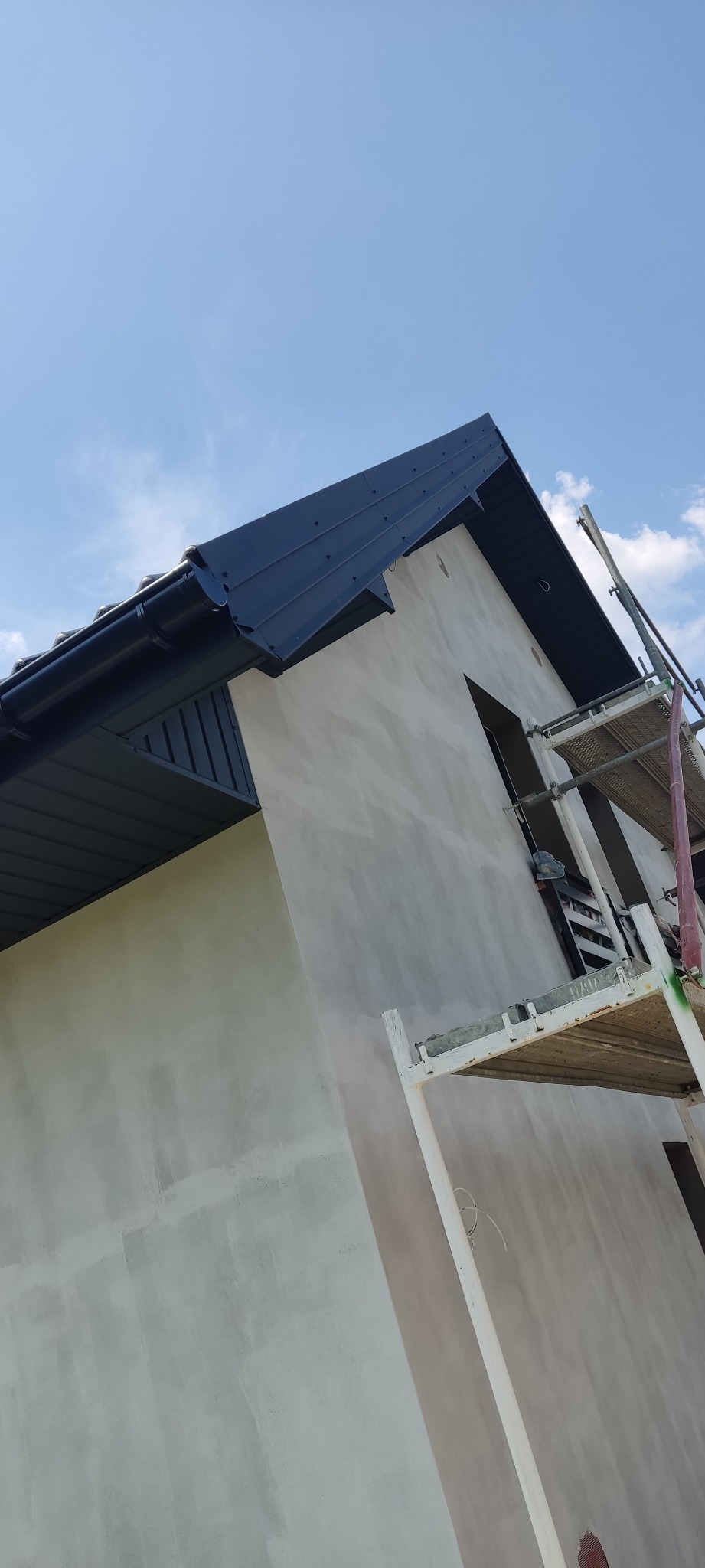 Nowy, ciemny dach z rynnami na tle jasnej elewacji domu w trakcie wykańczania, widoczne rusztowanie.