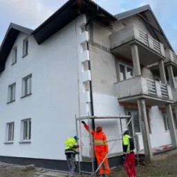 Dom w trakcie prac elewacyjnych: częściowo ocieplony styropianem, fragment pomalowany na biało, pracownicy w odblaskowych ubraniach montują rusztowanie.