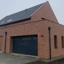 Nowoczesny dom jednorodzinny z ceglaną elewacją, grafitową stolarką okienną i bramą garażową, oraz dachem pokrytym grafitową dachówką. Widoczne orynnowanie w kolorze czarnym oraz wystające przewody...