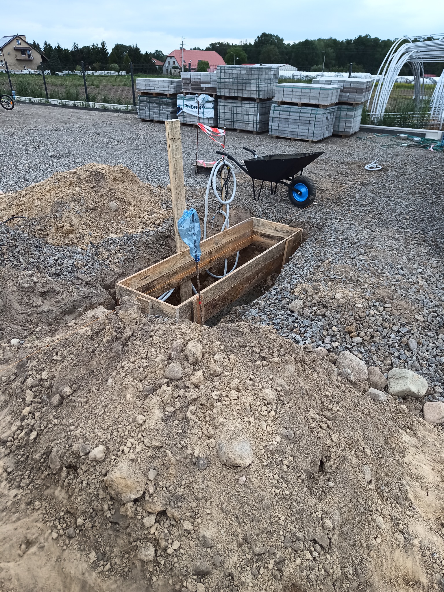 Wykop z drewnianą szalunką, widoczne rury wewnątrz, otoczony ziemią i gruzem, taczka z niebieskim kołem w tle, materiały budowlane w paletach.