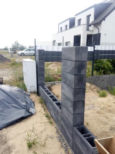 Wznoszenie fundamentu pod ogrodzenie z bloczków betonowych, widoczny fragment podmurówki z pustaków szalunkowych i słupki z bloczków, w tle siatka ogrodzeniowa i budynek mieszkalny.