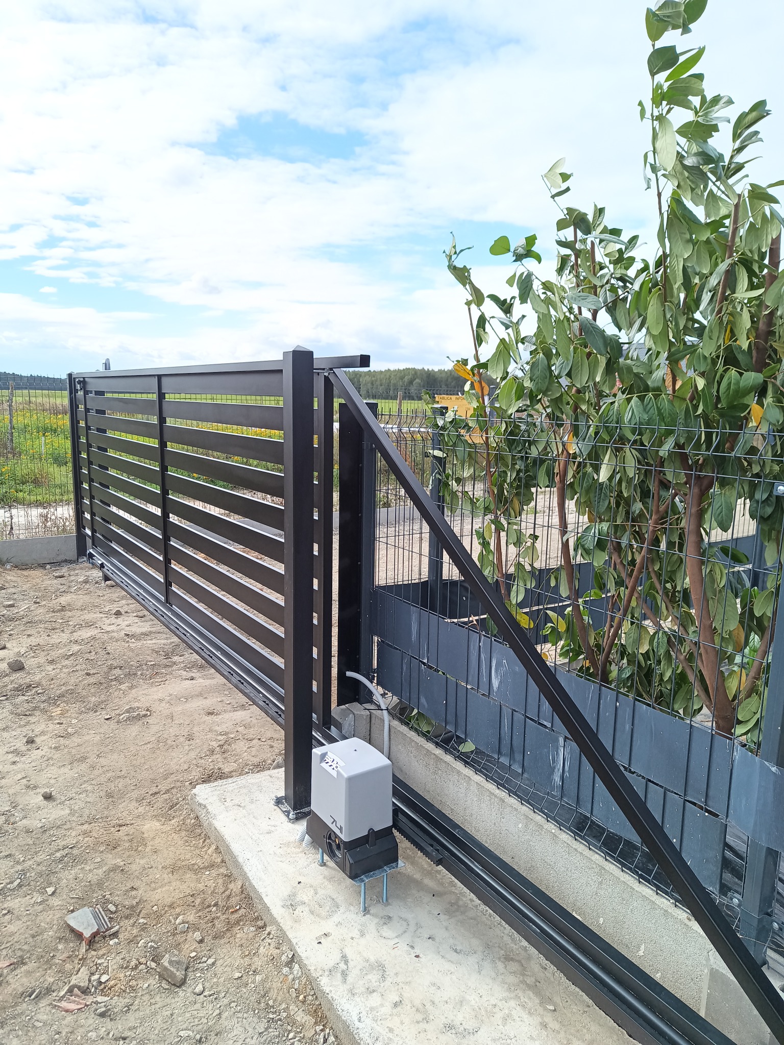 Nowoczesna, przesuwna brama wjazdowa z automatyką, widoczny napęd na betonowym fundamencie, obok siatka ogrodzeniowa i zieleń, na tle błękitne niebo.