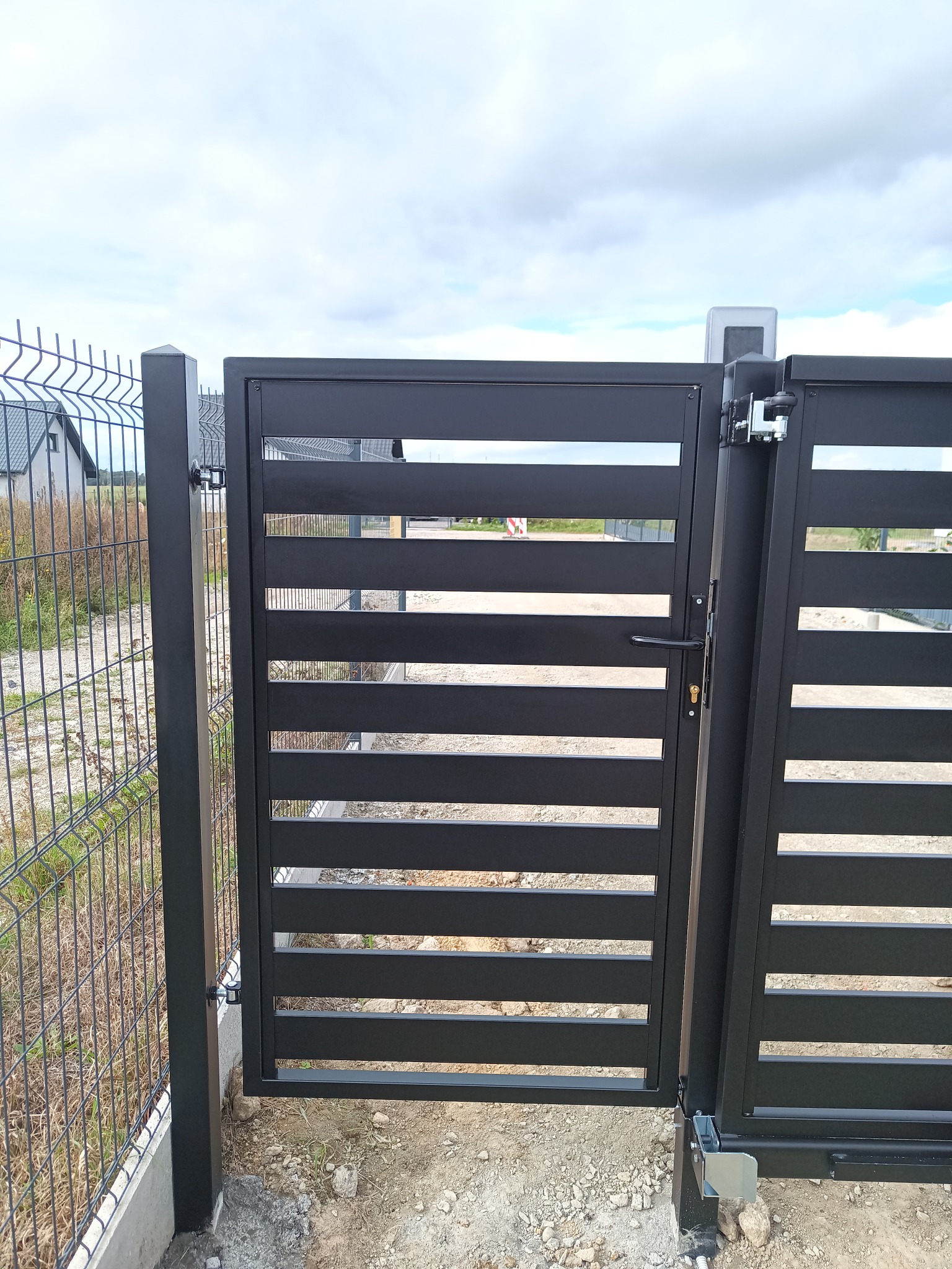 Nowoczesna, czarna brama wjazdowa z poziomymi panelami, widoczna automatyka i ogrodzenie z siatki w tle.