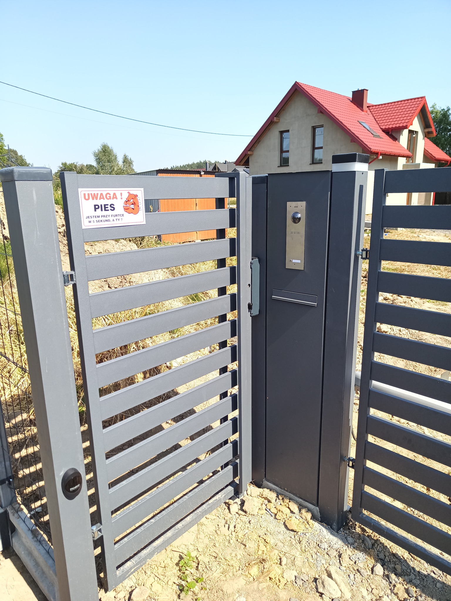 Otwarta szara furtka w ogrodzeniu panelowym z domofonem i skrzynką na listy, tabliczka ostrzegawcza 'Uwaga! Pies' po lewej stronie