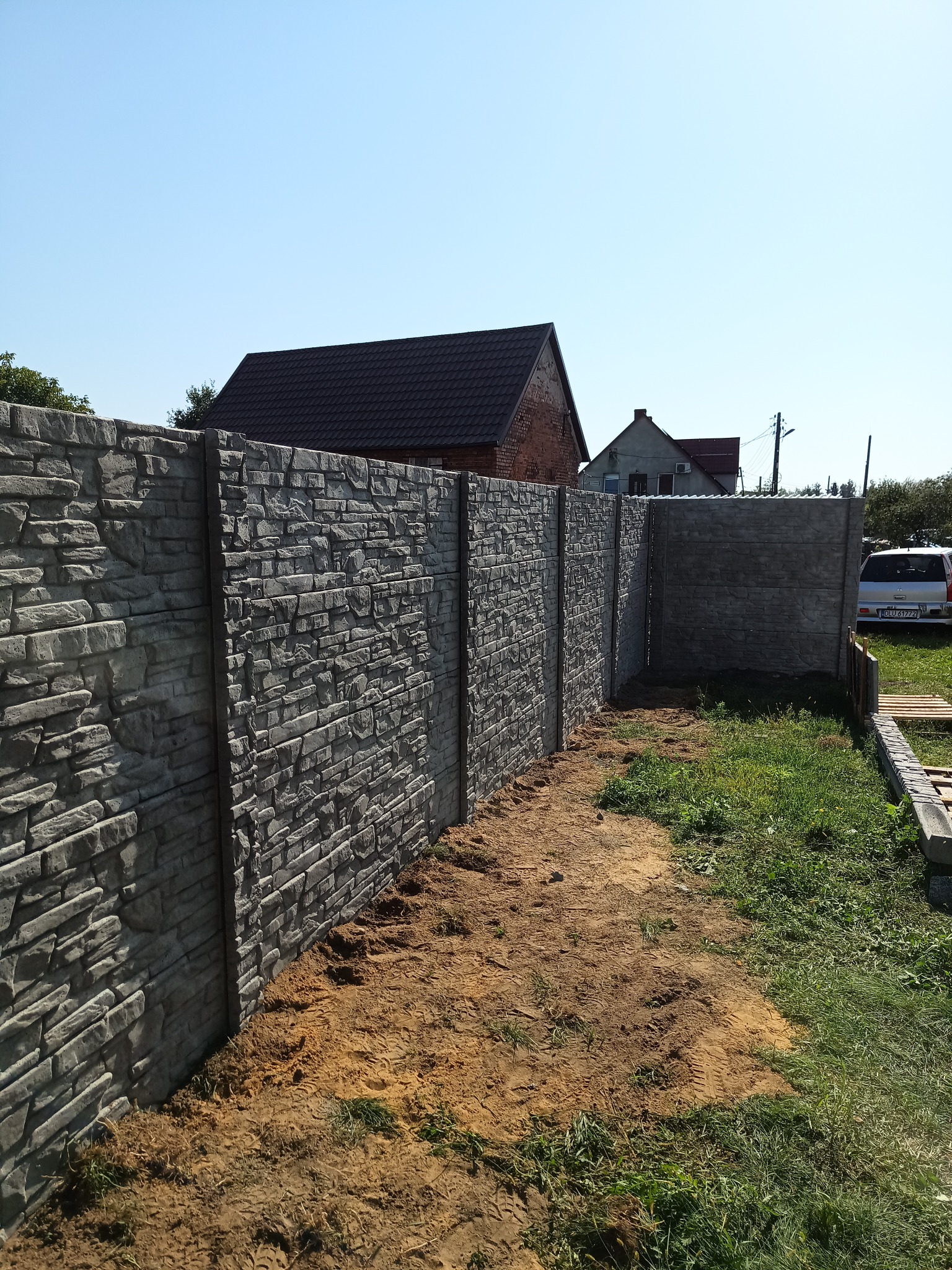 Szary, betonowy płot imitujący kamień w trakcie montażu, z widoczną ziemią i trawą, w tle domy i błękitne niebo.