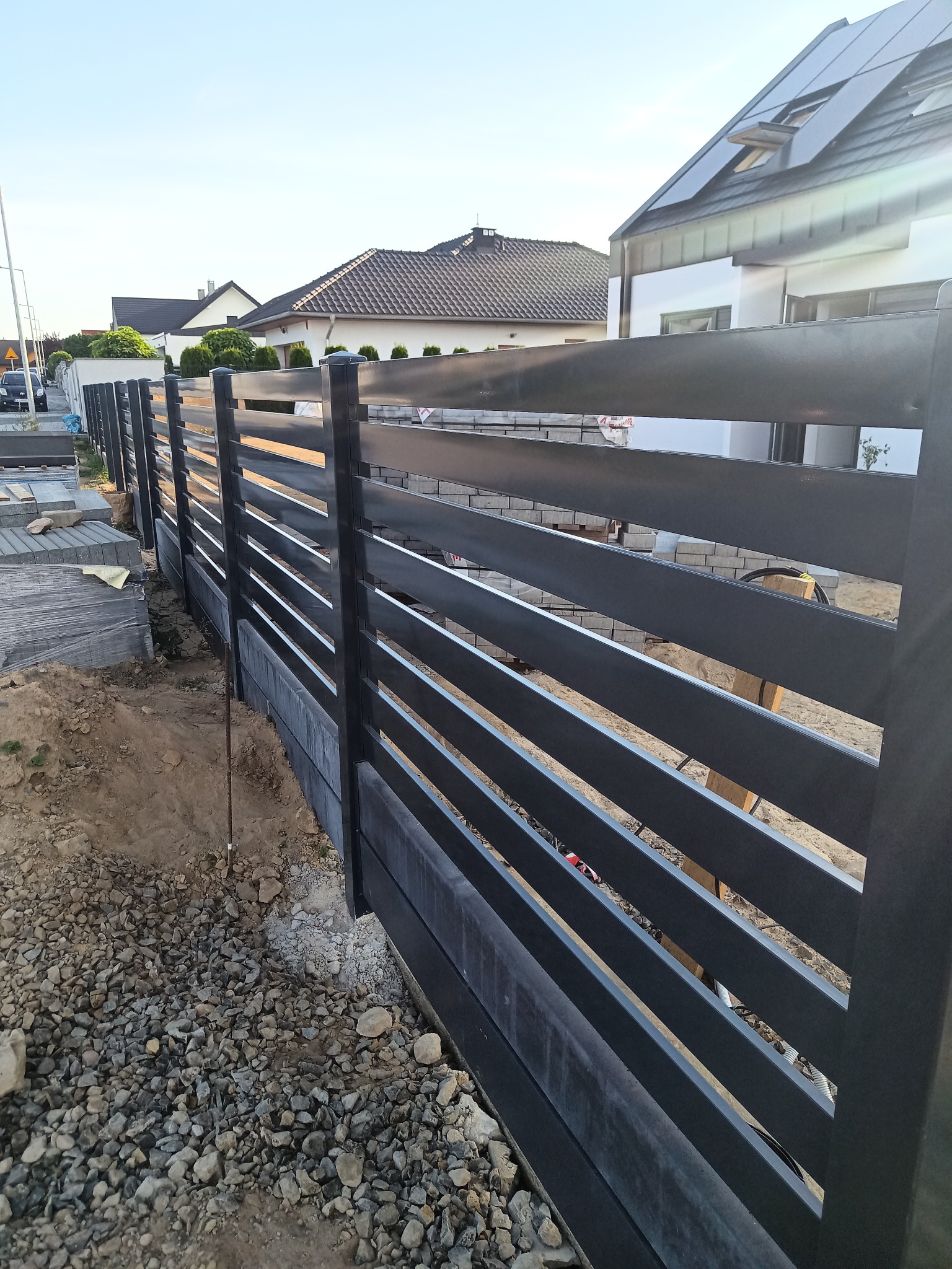 Montaż nowoczesnego ogrodzenia panelowego z poziomymi przęsłami w kolorze grafitowym, widoczne przygotowane podłoże z kamieni i kostki brukowej w tle.