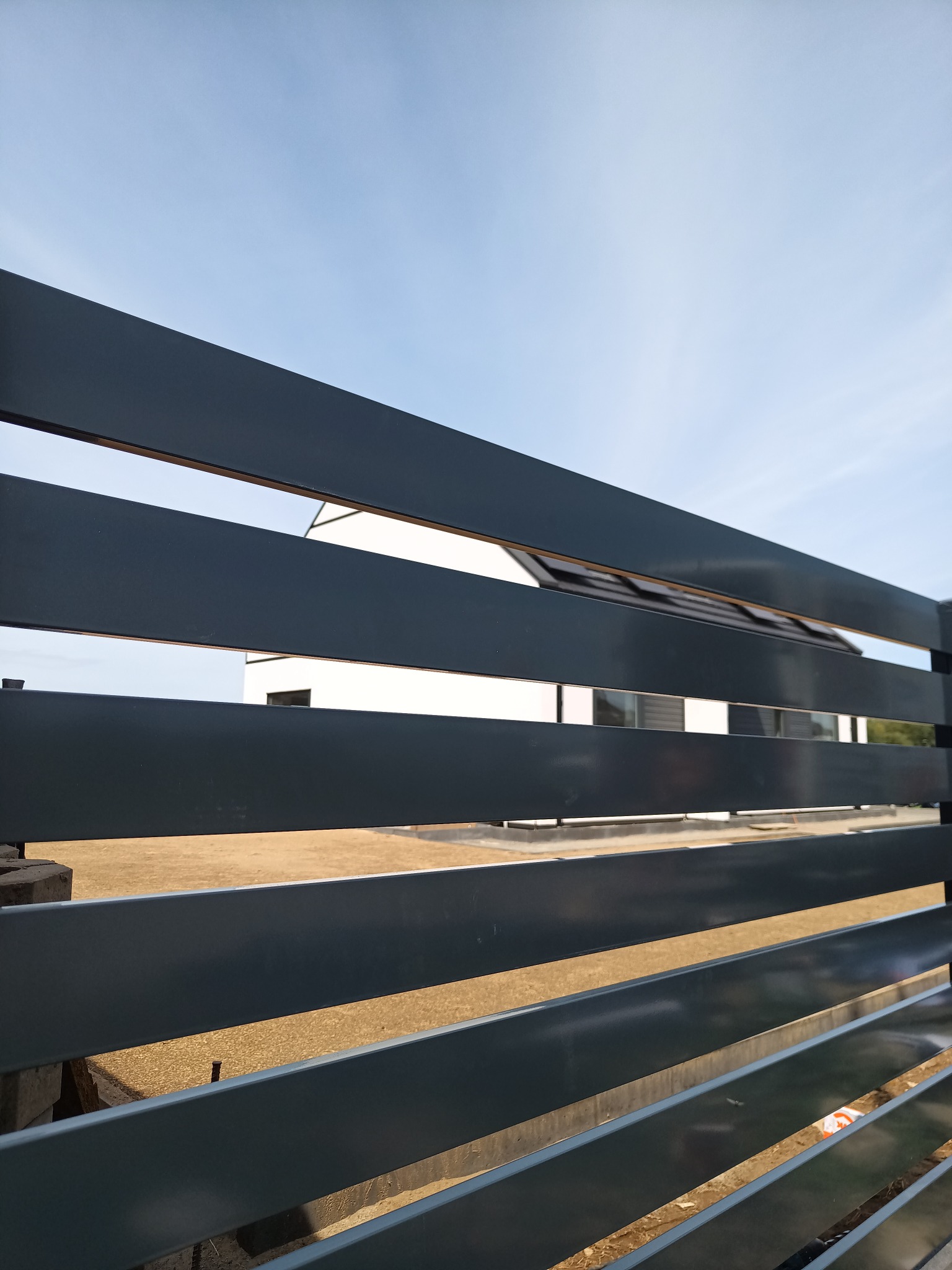 Szare, poziome przęsła ogrodzenia z widokiem na biały dom z ciemnym dachem na tle jasnego nieba.