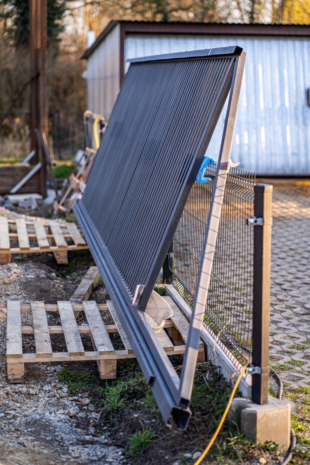 Panel ogrodzeniowy oparty o istniejące ogrodzenie i palety, przygotowany do montażu. Widoczna metalowa konstrukcja i fragment trawnika.