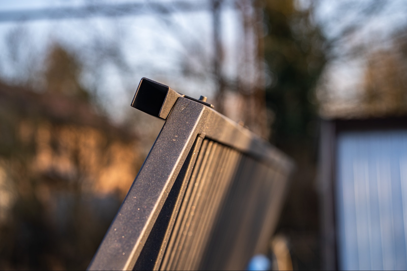 Metalowy panel ogrodzeniowy w zbliżeniu, widoczne detale konstrukcji i łączenia elementów, tło rozmyte, ciepłe światło podkreśla fakturę materiału.