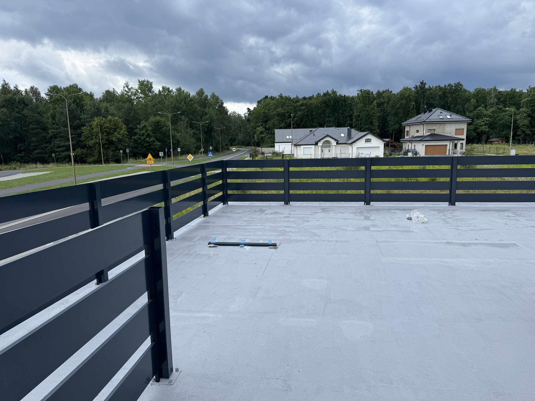 Nowoczesne, poziome ogrodzenie panelowe w kolorze antracytowym, z widocznym mocowaniem. W tle zabudowa jednorodzinna, zieleń i pochmurne niebo. Minimalistyczny design.