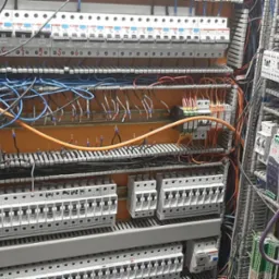 Rozdzielnia elektryczna z widocznymi bezpiecznikami, przewodami w różnych kolorach (pomarańczowy, niebieski, czarny), oraz elementami automatyki, częściowo zakryta kartką z notatkami.