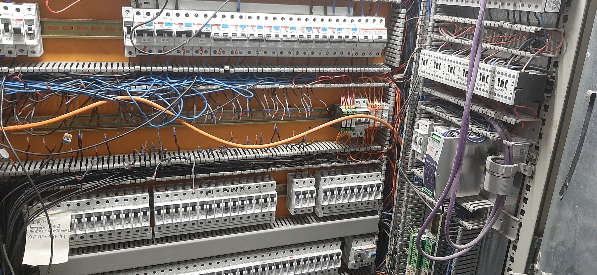 Rozdzielnia elektryczna z widocznymi bezpiecznikami, przewodami w różnych kolorach (pomarańczowy, niebieski, czarny), oraz elementami automatyki, częściowo zakryta kartką z notatkami.