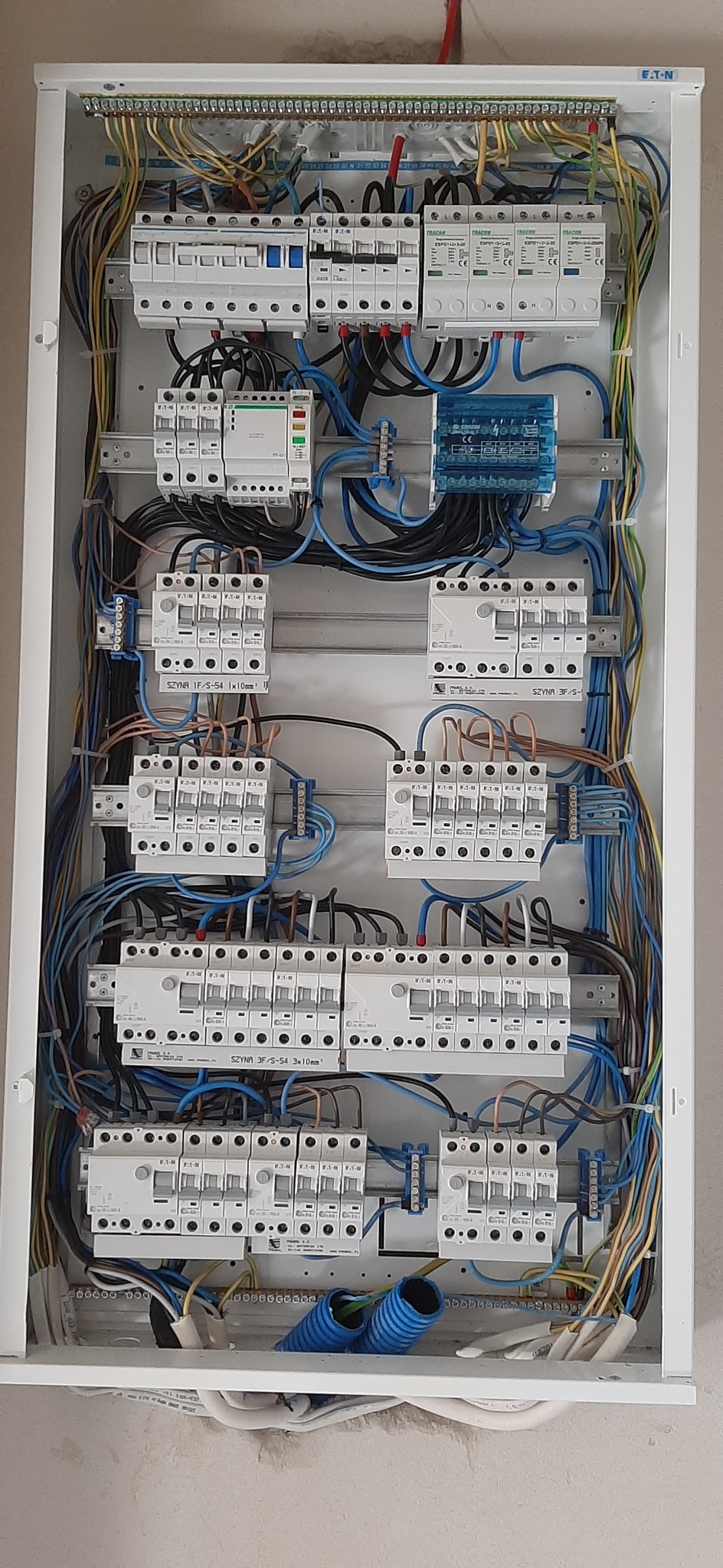 Rozdzielnica elektryczna z zainstalowanymi bezpiecznikami, przewodami i modułami, widoczne niebieskie rury karbowane w dolnej części skrzynki.