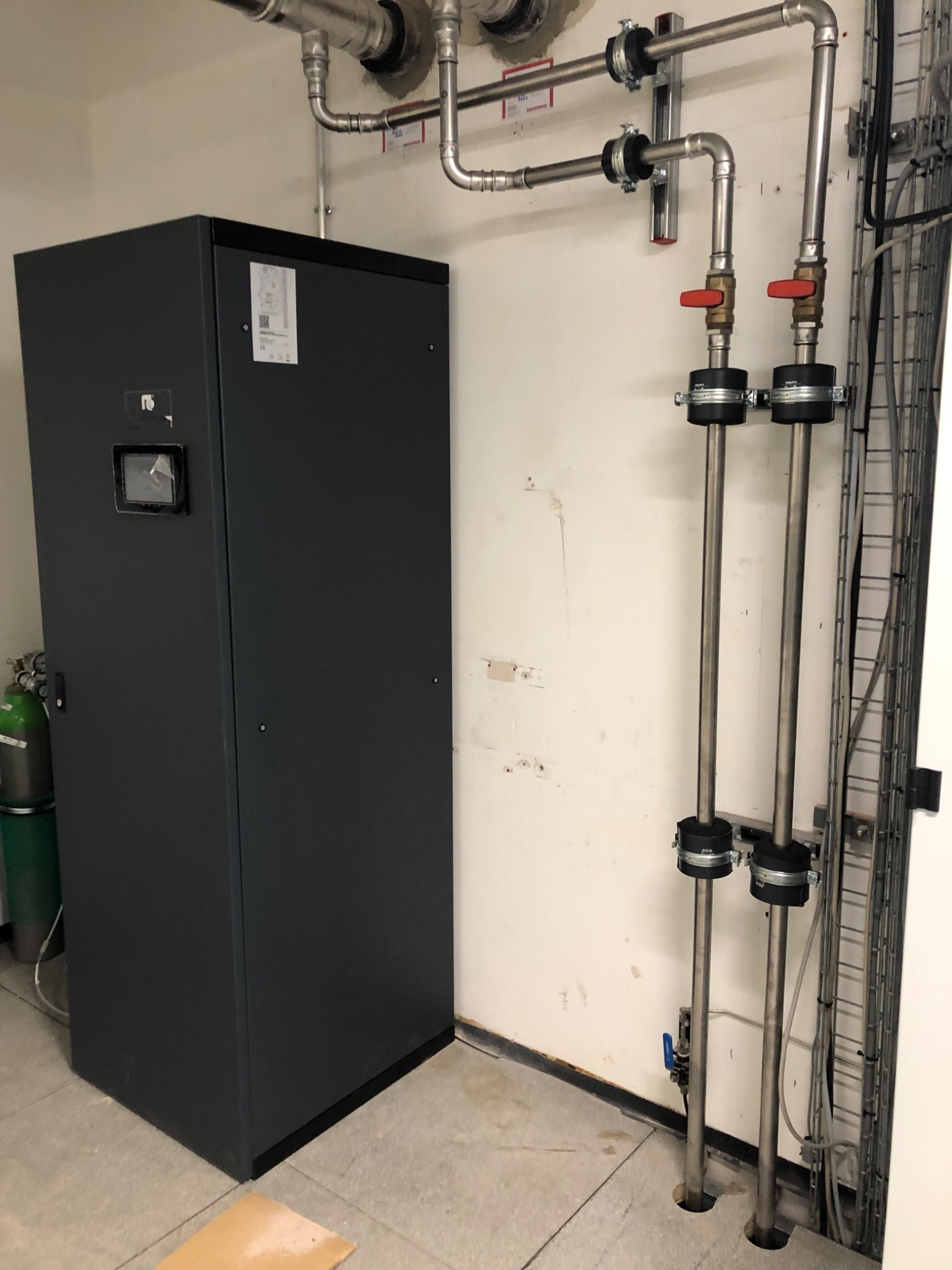 Szara, metalowa szafa sterownicza z wyświetlaczem w kotłowni, widoczne rury instalacji grzewczej z zaworami i mocowaniami do ściany.