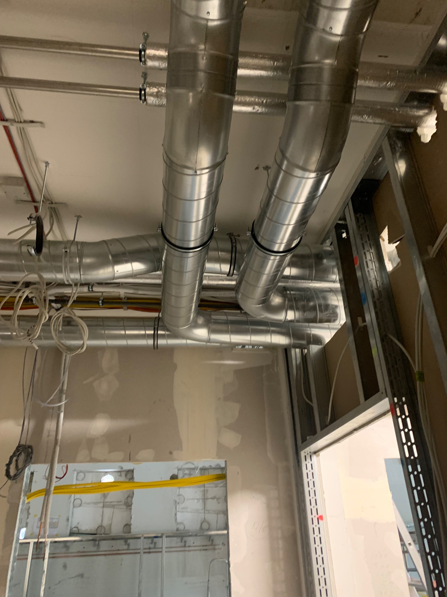 Widok na sufit w trakcie montażu instalacji wentylacyjnej, z widocznymi metalowymi rurami wentylacyjnymi, przewodami elektrycznymi i elementami konstrukcyjnymi ścian.