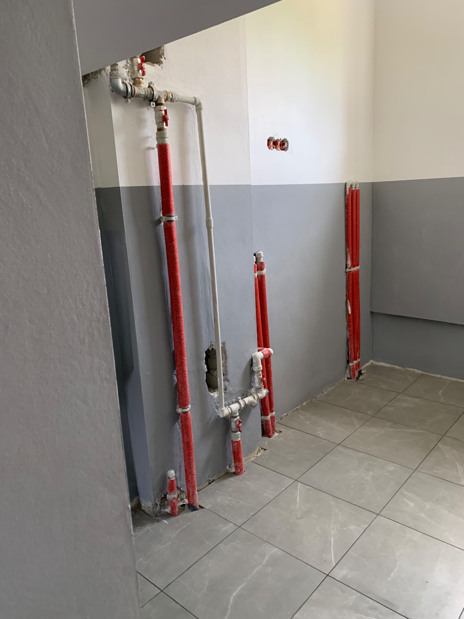 Wnętrze łazienki w trakcie remontu z widocznymi rurami instalacji wodnej i grzewczej, częściowo ukrytymi w ścianie, z widocznymi przyłączami i zaworami, oraz fragmentem nowej podłogi z płytek.