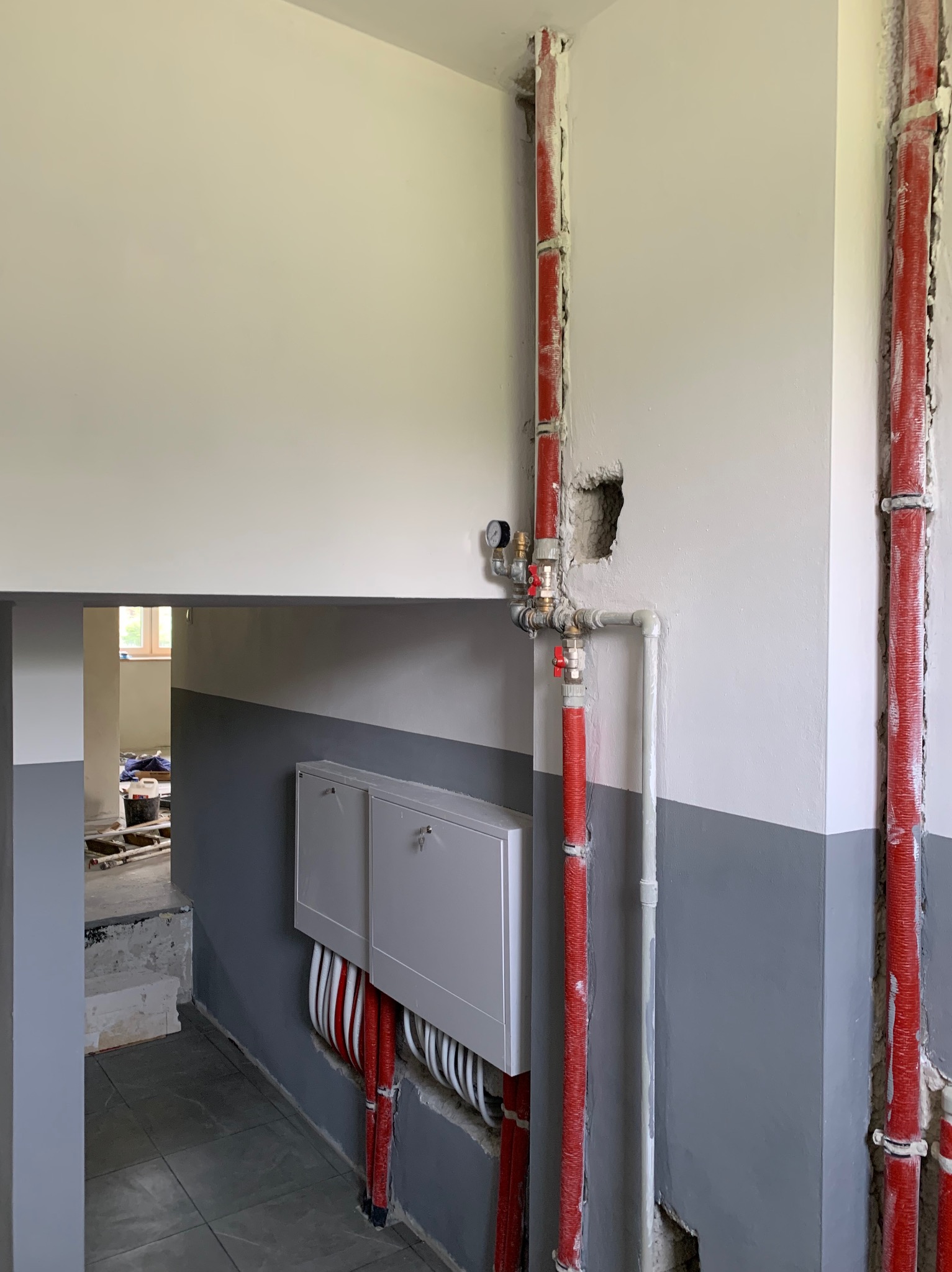 Instalacja rur grzewczych w nowym budynku, widoczne czerwone rury z izolacją, biała skrzynka rozdzielcza z rurami podłogowymi, ściany pomalowane na biało i szaro, częściowo zakryte niedoskonałości...