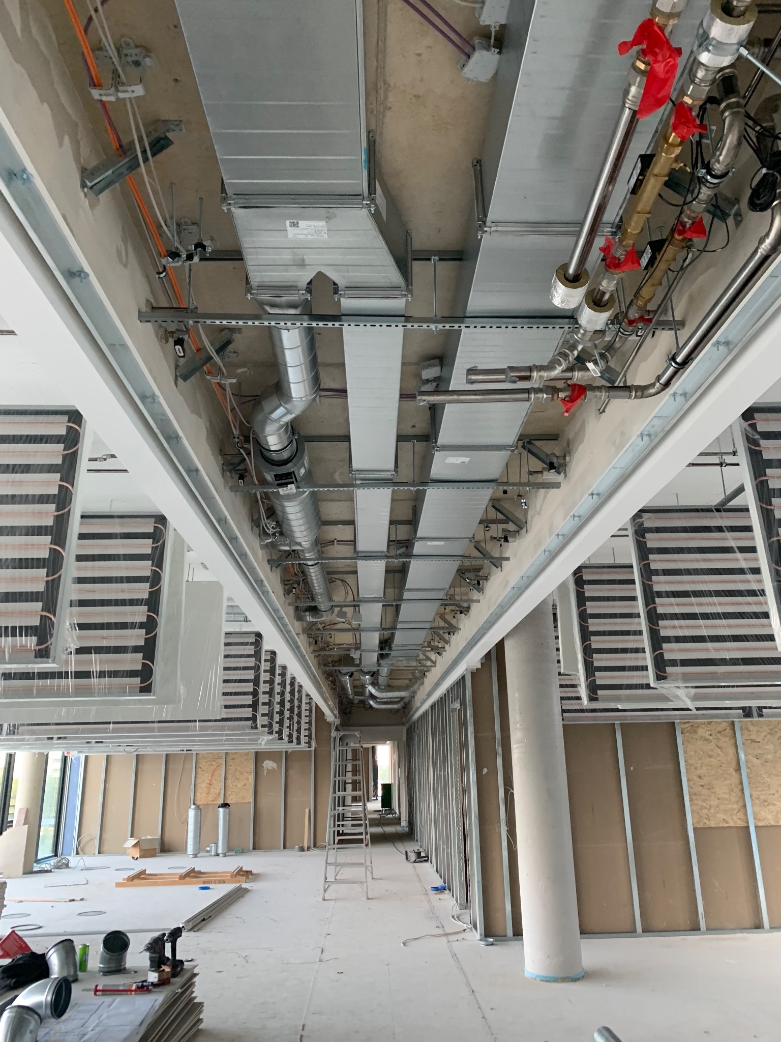 Widok z dołu na sufit w trakcie montażu instalacji: rury, kanały wentylacyjne, elementy ogrzewania podłogowego, drabina w tle.