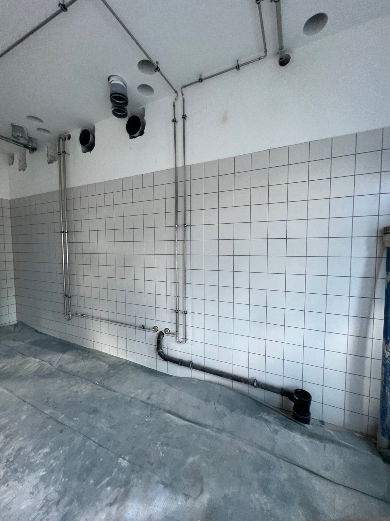 Wnętrze w trakcie prac hydraulicznych: widoczne rury ze stali nierdzewnej przymocowane do ściany z białych płytek, czarne rury kanalizacyjne przy podłodze zabezpieczonej folią, oraz otwory w suficie.