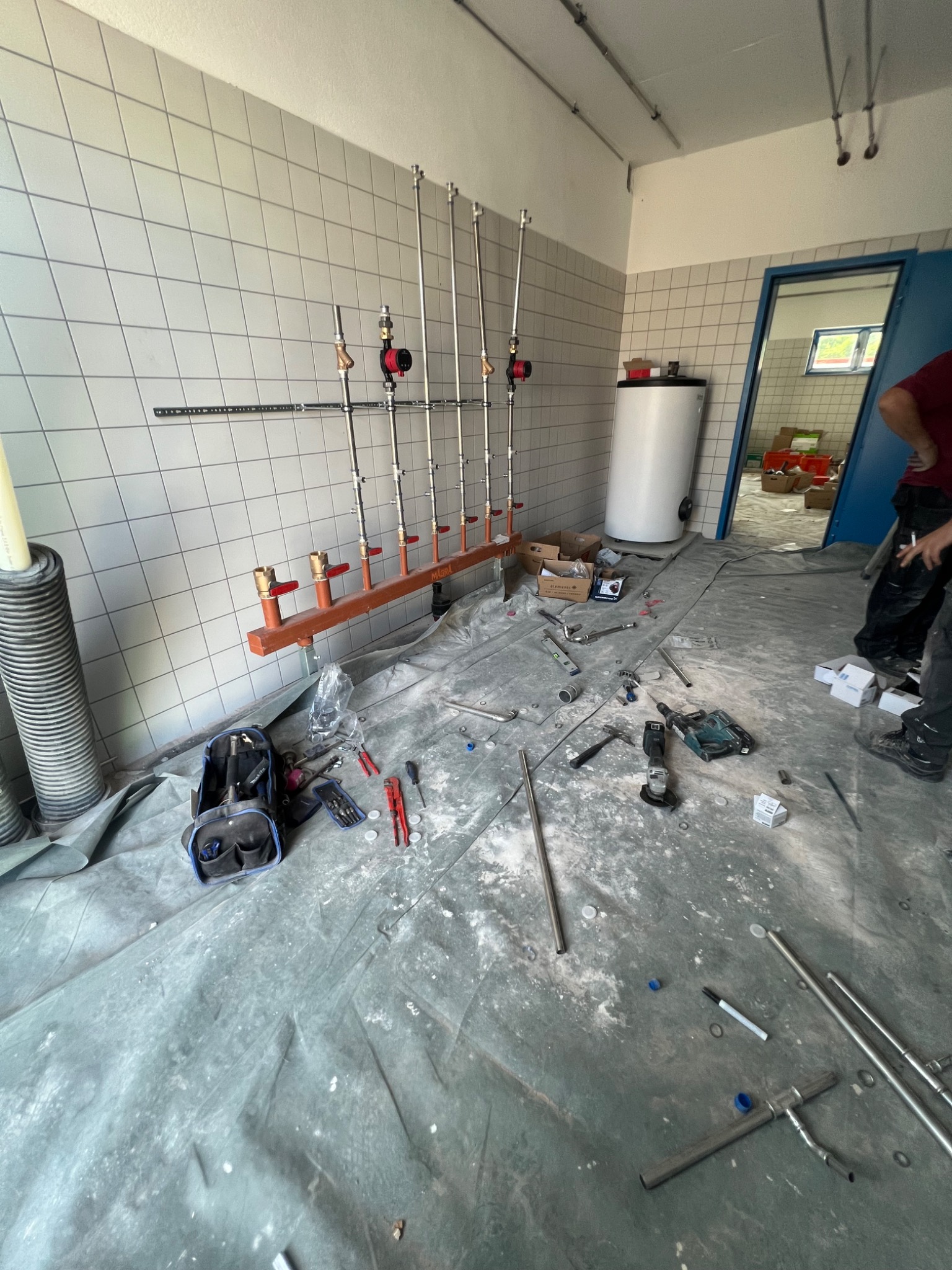 Montaż instalacji wodnej: rury pionowe z czerwonymi zaworami i brązową belką mocującą na tle białych płytek, narzędzia i materiały rozłożone na folii ochronnej, w tle biały bojler i otwarte drzwi.