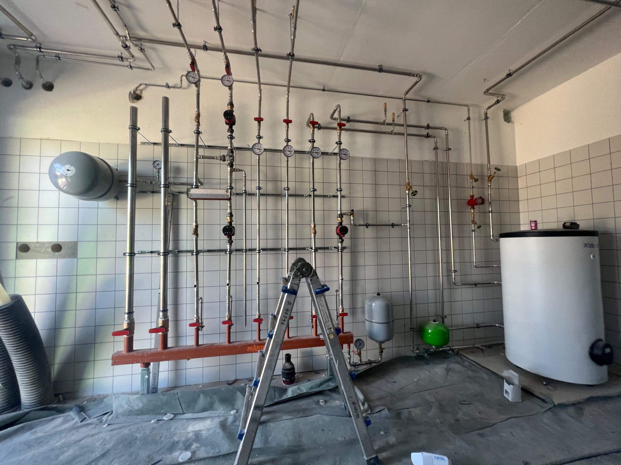 Montaż instalacji grzewczej: widoczne liczne rury, zawory, manometry, zbiornik wyrównawczy oraz drabina monterska na tle ściany wyłożonej kafelkami.