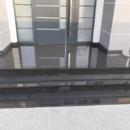 Trzy stopnie wejściowe z czarnego, polerowanego kamienia prowadzące do nowoczesnych drzwi z elementami szkła i szarymi ramami, z widocznym fragmentem chodnika z jasnoszarej kostki brukowej.