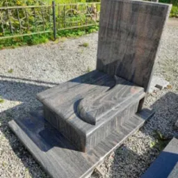 Wypolerowany, dwupoziomowy nagrobek z ciemnego, wzorzystego granitu na żwirowym podłożu, z niskim metalowym ogrodzeniem w tle.