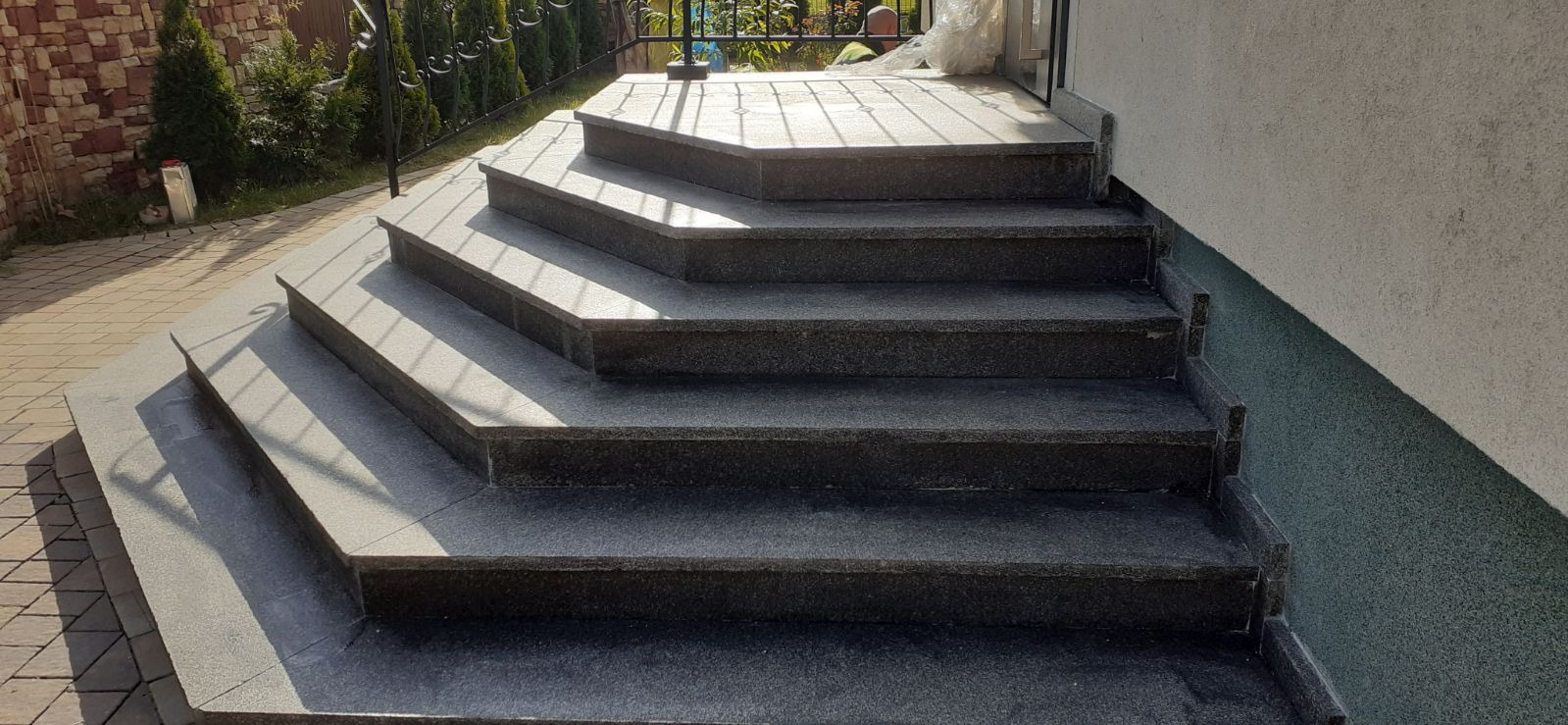 Zewnętrzne schody wejściowe z ciemnego kamienia, prowadzące do drzwi. Widoczne ogrodzenie i fragment ściany budynku.