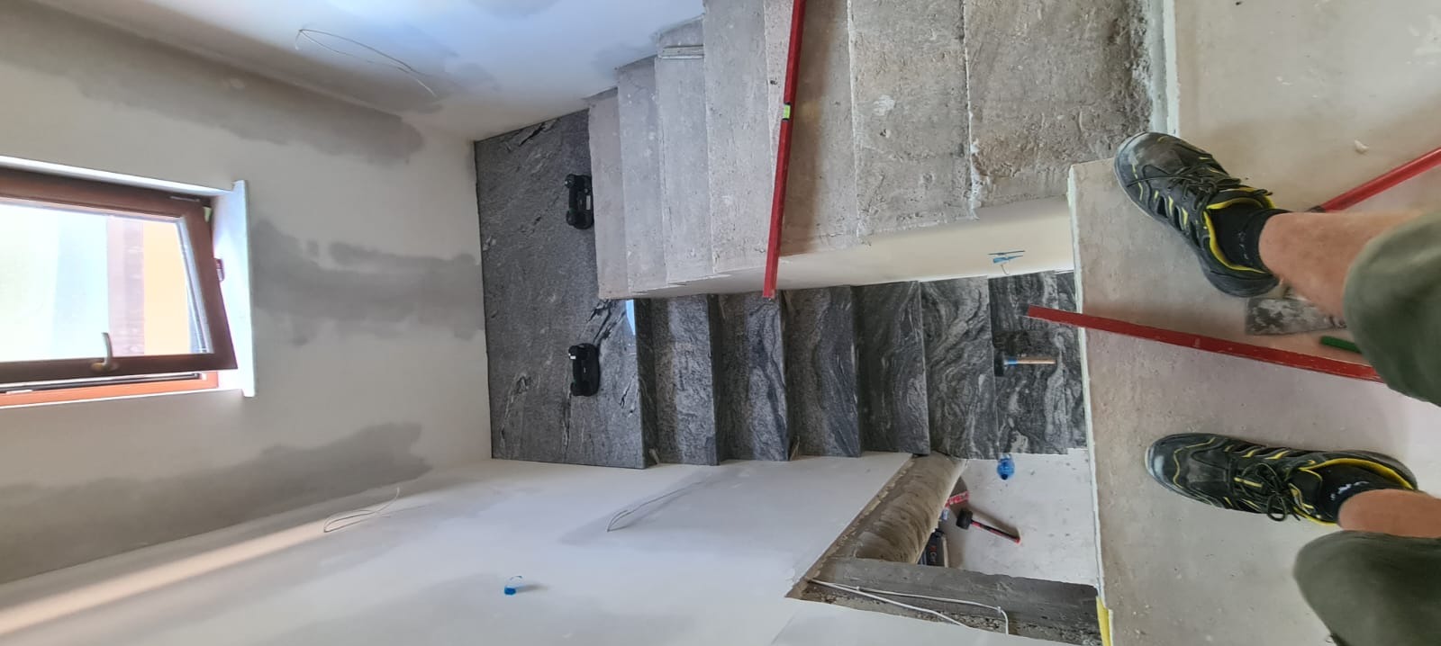 Montaż stopni schodowych z szarego kamienia z wyraźnym wzorem żyłkowania, widok z góry z fragmentem osoby w butach roboczych, poziomica i młotek w kadrze, surowy stan wykończenia wnętrza.