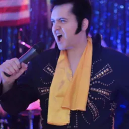 Mężczyzna w stroju imitującym Elvisa Presleya, śpiewający do mikrofonu z ekspresją, w tle częściowo widoczna flaga USA i dekoracje świetlne.