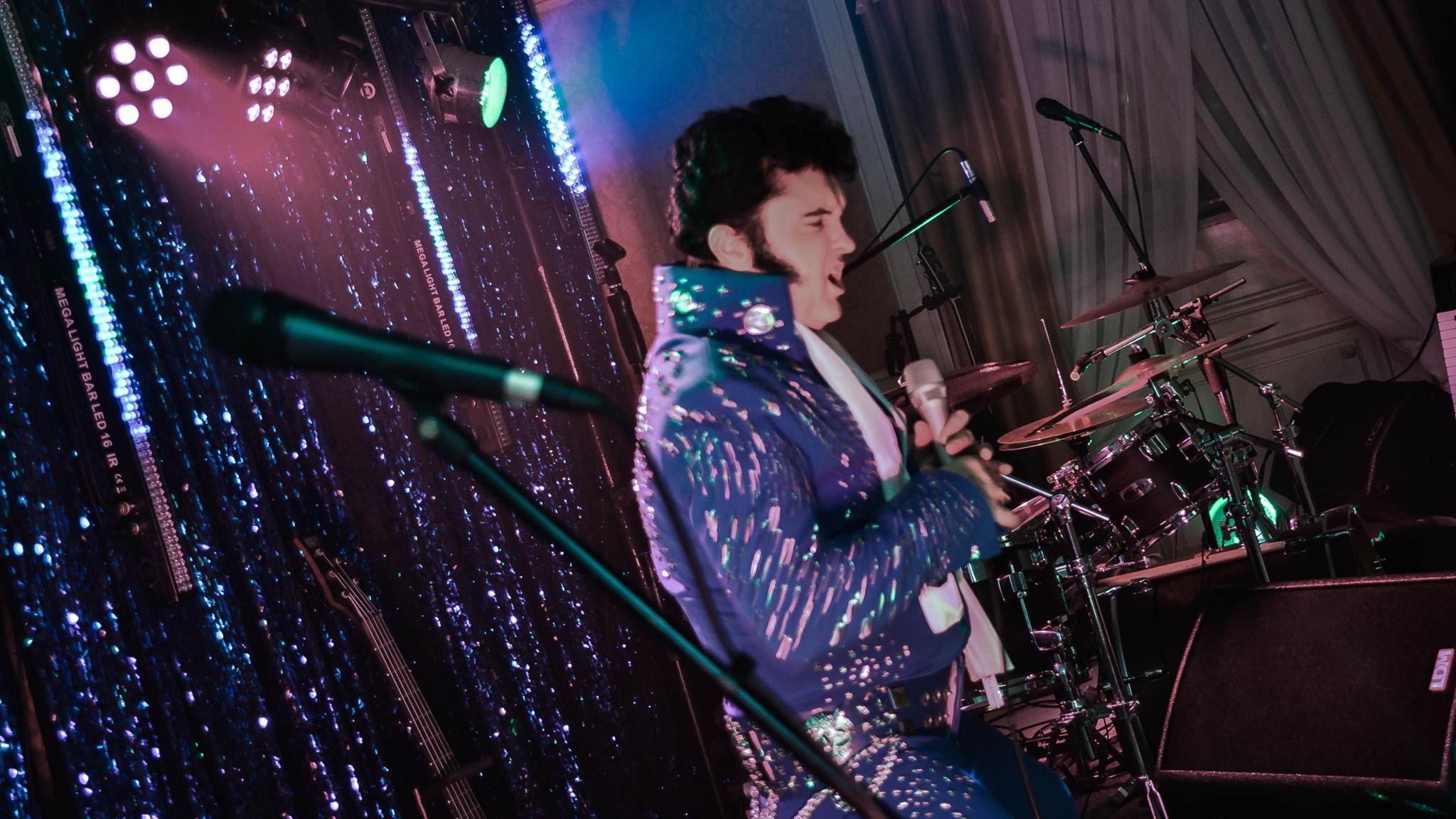 Mężczyzna w błyszczącym, niebieskim kostiumie w stylu Elvisa Presleya śpiewa do mikrofonu na scenie z perkusją i oświetleniem LED w tle.