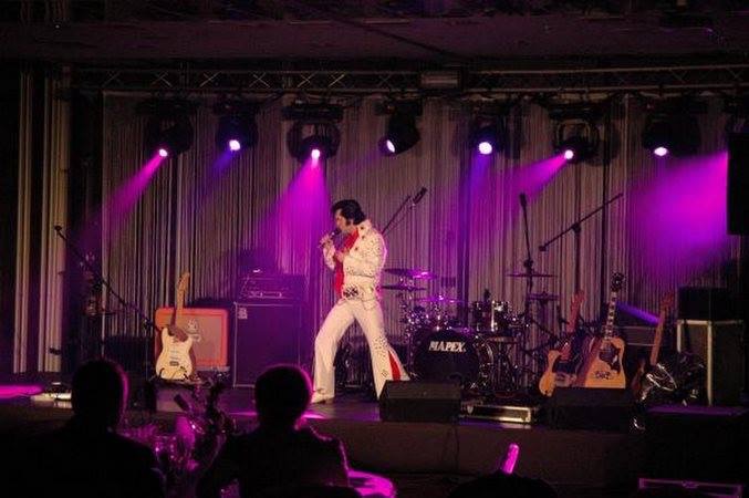 Elvis Presley impersonator performing on stage with a full band, under purple spotlights, in front of a curtain backdrop.