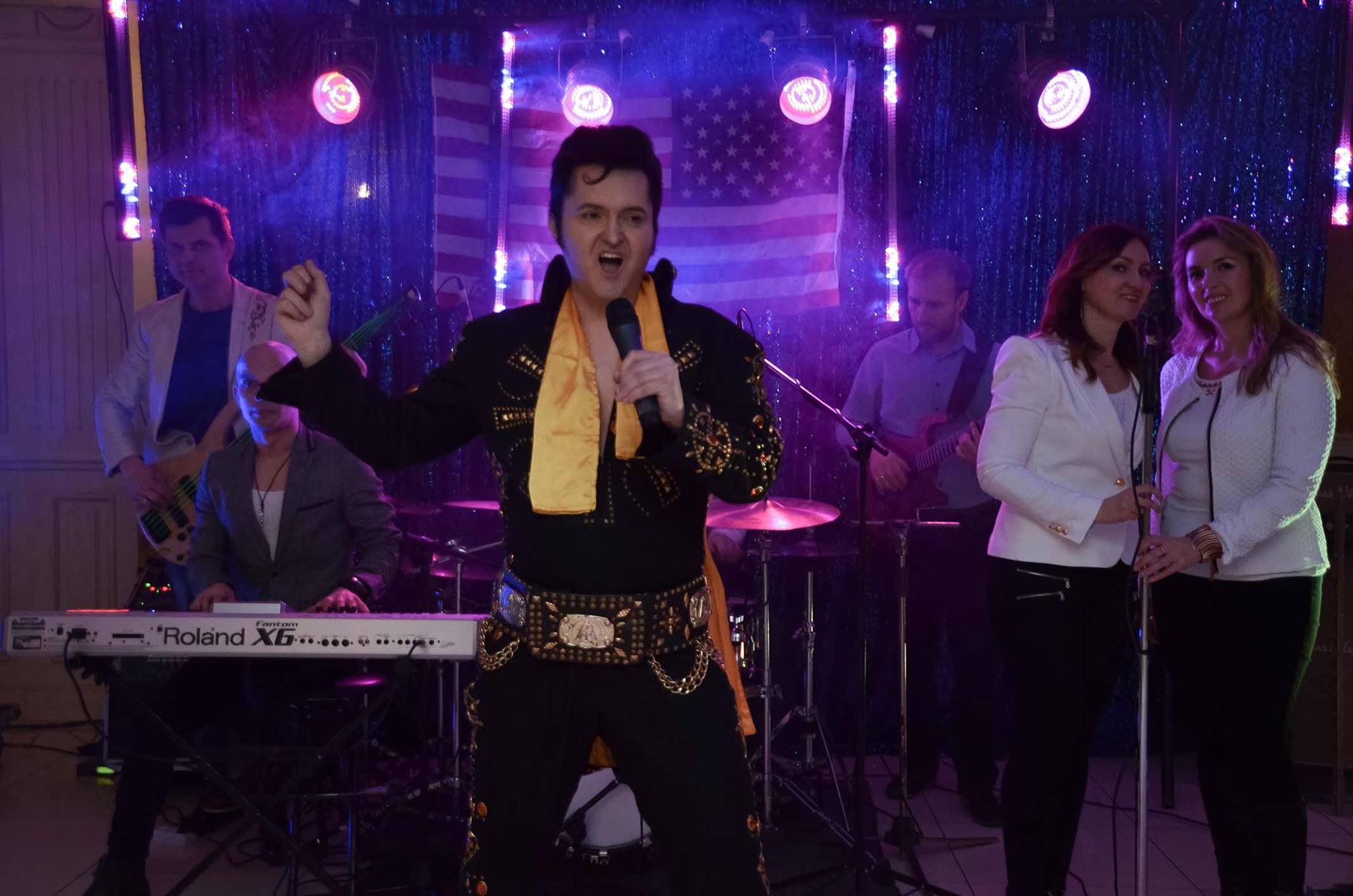 Wokalista przebrany za Elvisa Presleya śpiewa do mikrofonu na scenie z zespołem, przed amerykańską flagą i dekoracjami świetlnymi.