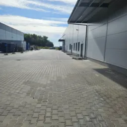 Szara kostka brukowa ułożona przed budynkiem magazynowym w słoneczny dzień. Widok z perspektywy pieszego.