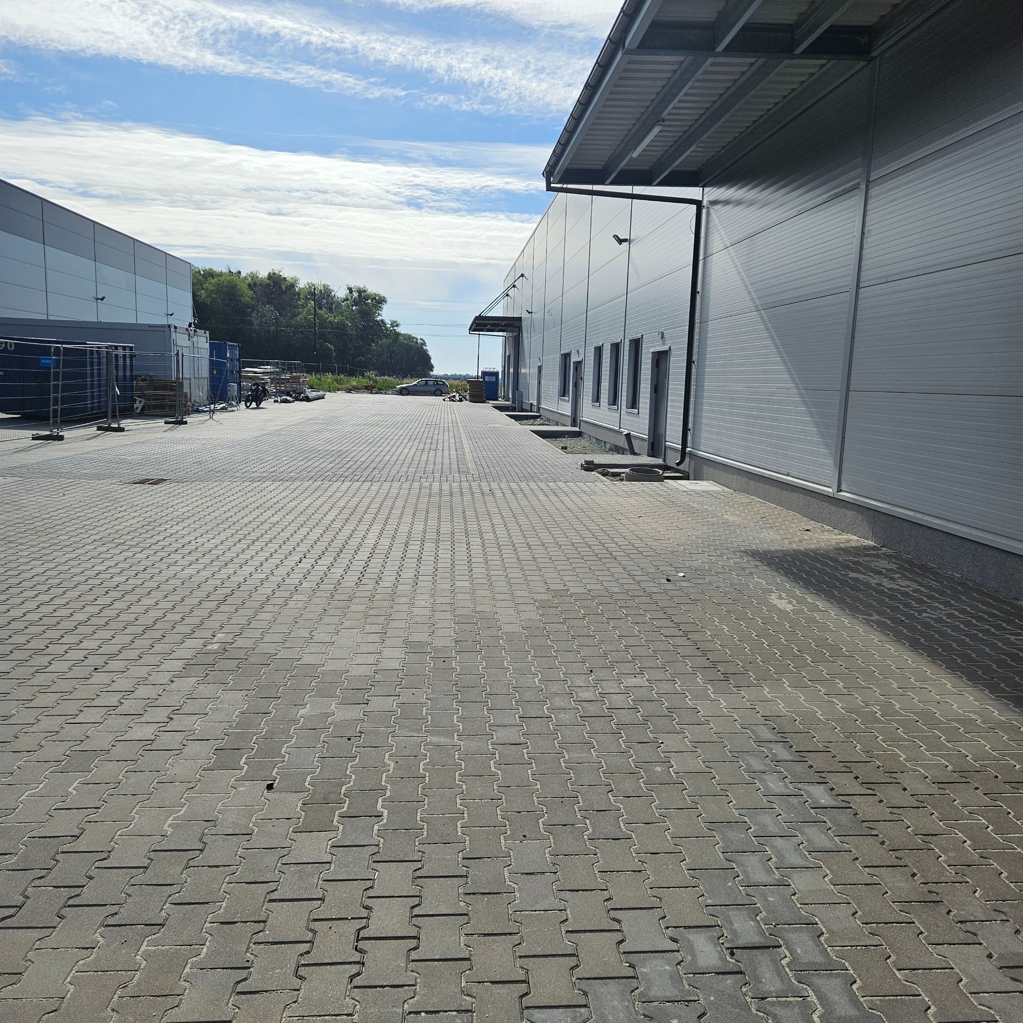 Szara kostka brukowa ułożona przed budynkiem magazynowym w słoneczny dzień. Widok z perspektywy pieszego.