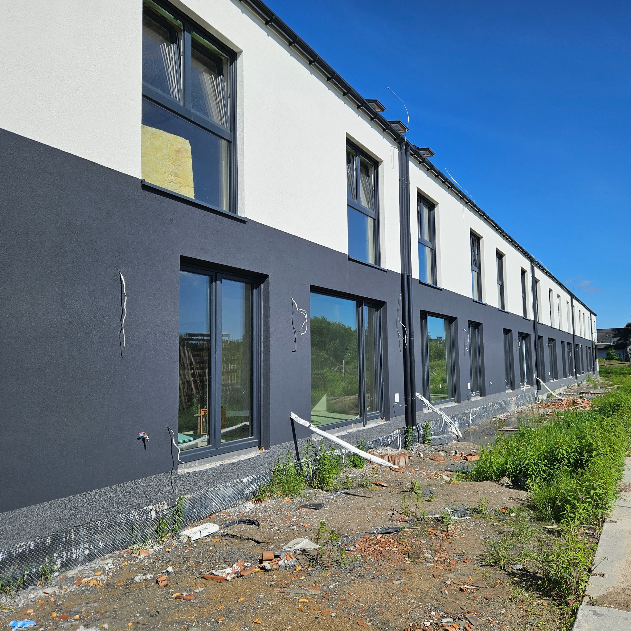 Elewacja budynku po ociepleniu, z widocznymi oknami PCV i fragmentem styropianu w Ludwinowie. Dwukolorowa fasada: szary dół i biała góra. Prace wykończeniowe.