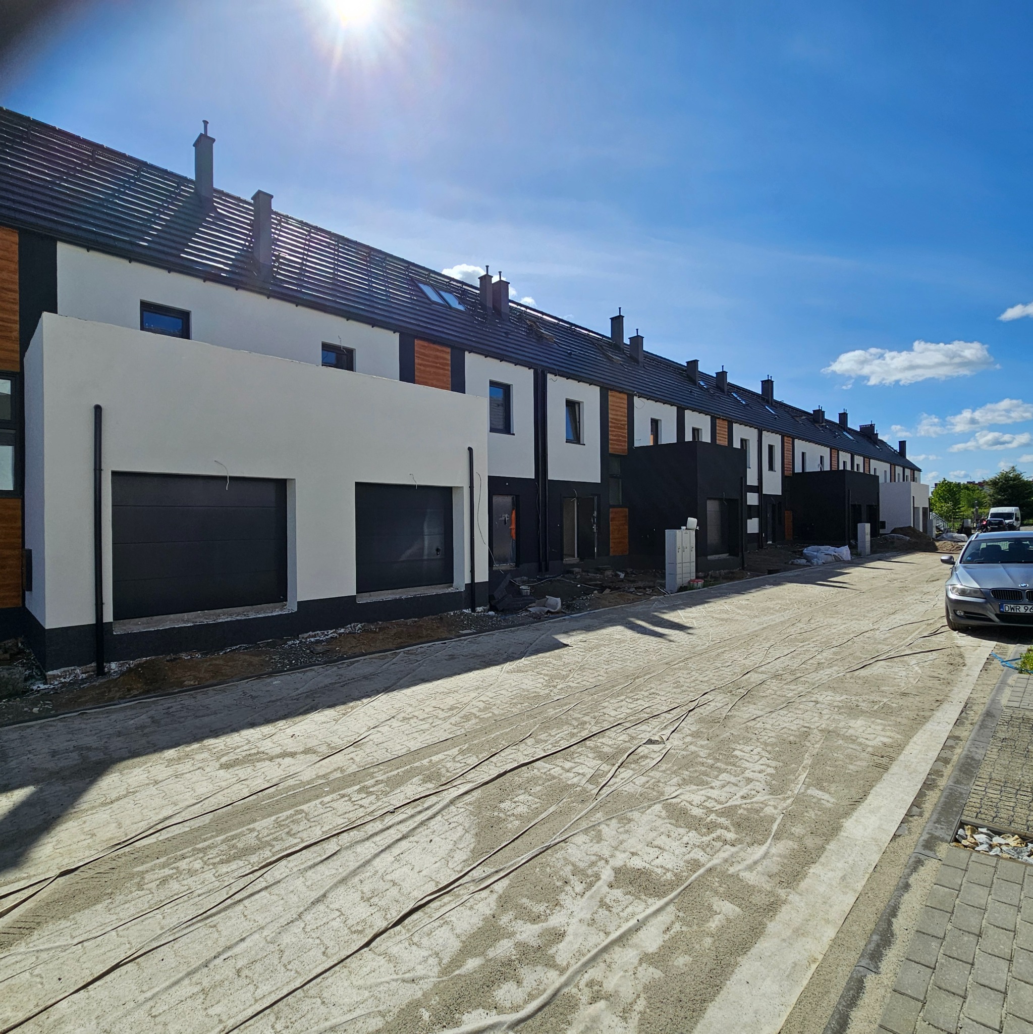 Nowoczesne szeregówki w trakcie wykończenia, widok z ulicy z przygotowanym podłożem, jasne elewacje z elementami drewna i czerni, błękitne niebo.