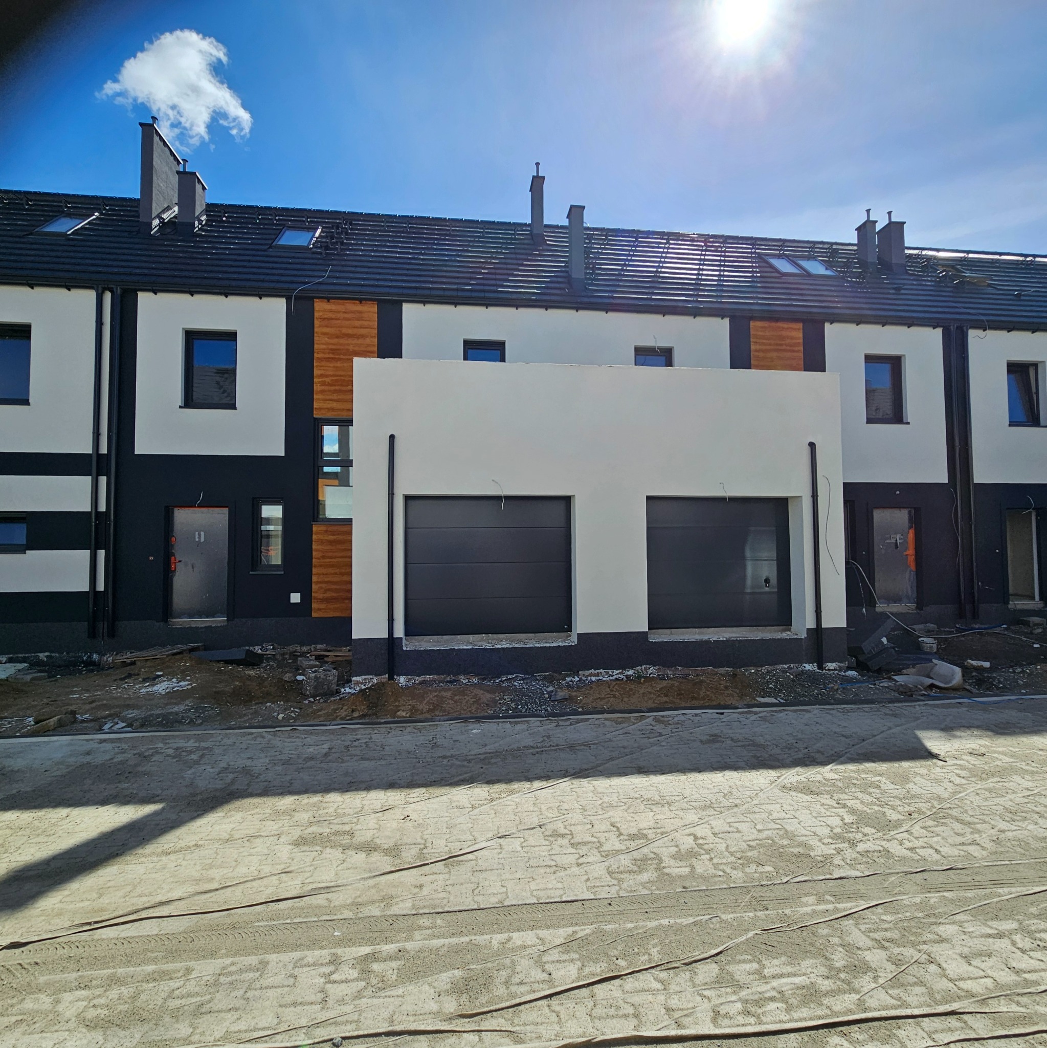 Nowoczesny szereg domów w trakcie wykończenia, z czarnymi bramami garażowymi i elementami drewnianej elewacji. Widoczne prace brukarskie przed budynkami w słoneczny dzień.