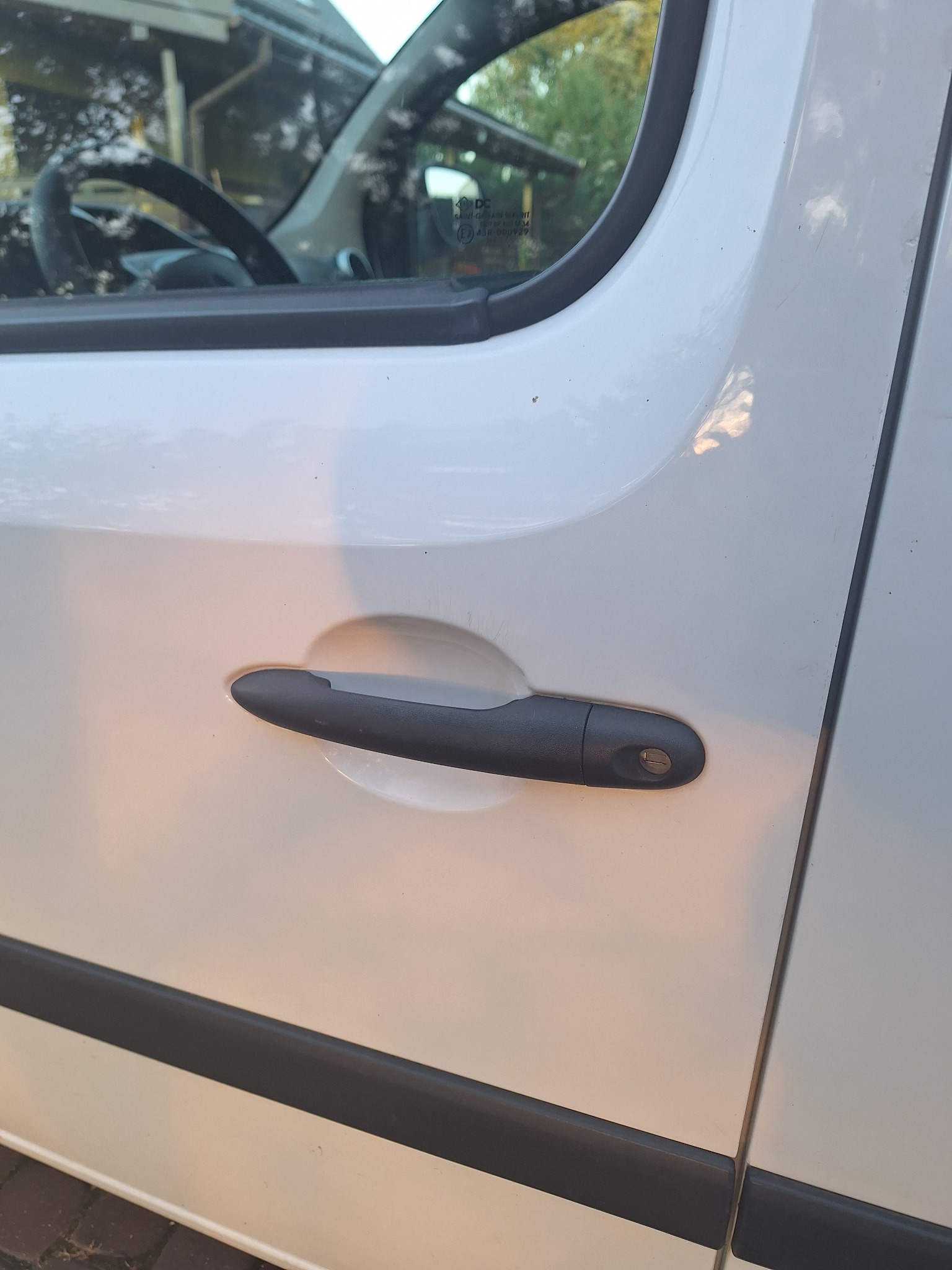 Czarny uchwyt drzwi bocznych białego samochodu dostawczego, widoczne zarysowania na lakierze wokół zamka.