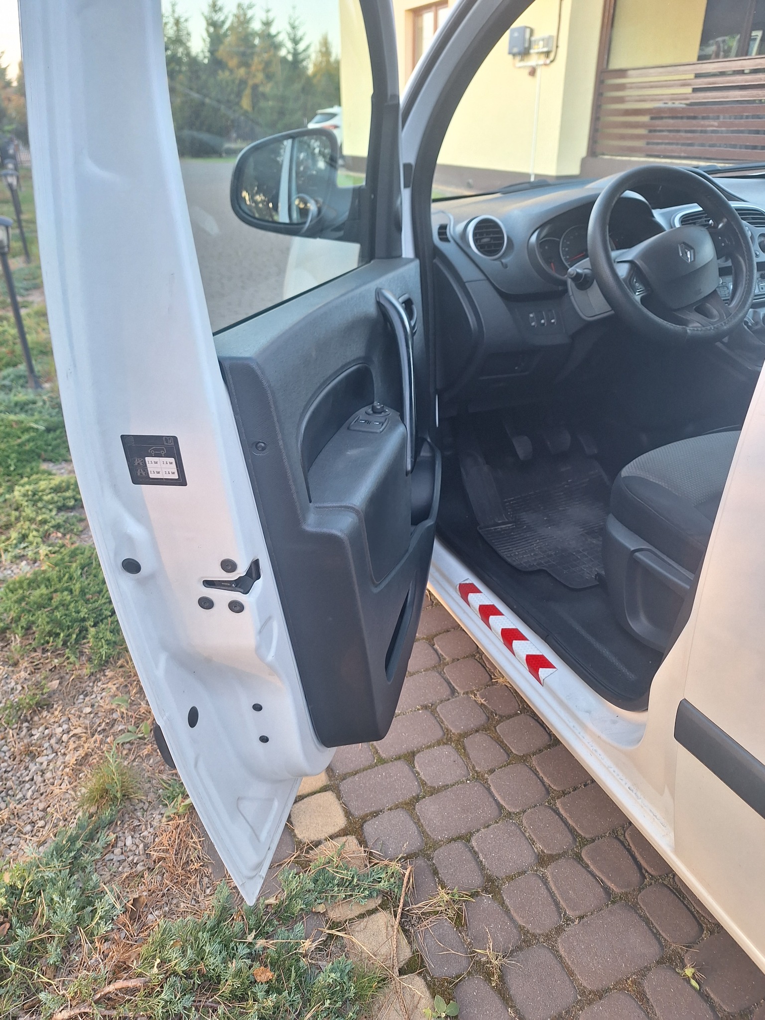 Wnętrze białego samochodu dostawczego z otwartymi drzwiami kierowcy, widoczne elementy deski rozdzielczej, kierownica, dywaniki oraz naklejki ostrzegawcze na drzwiach.