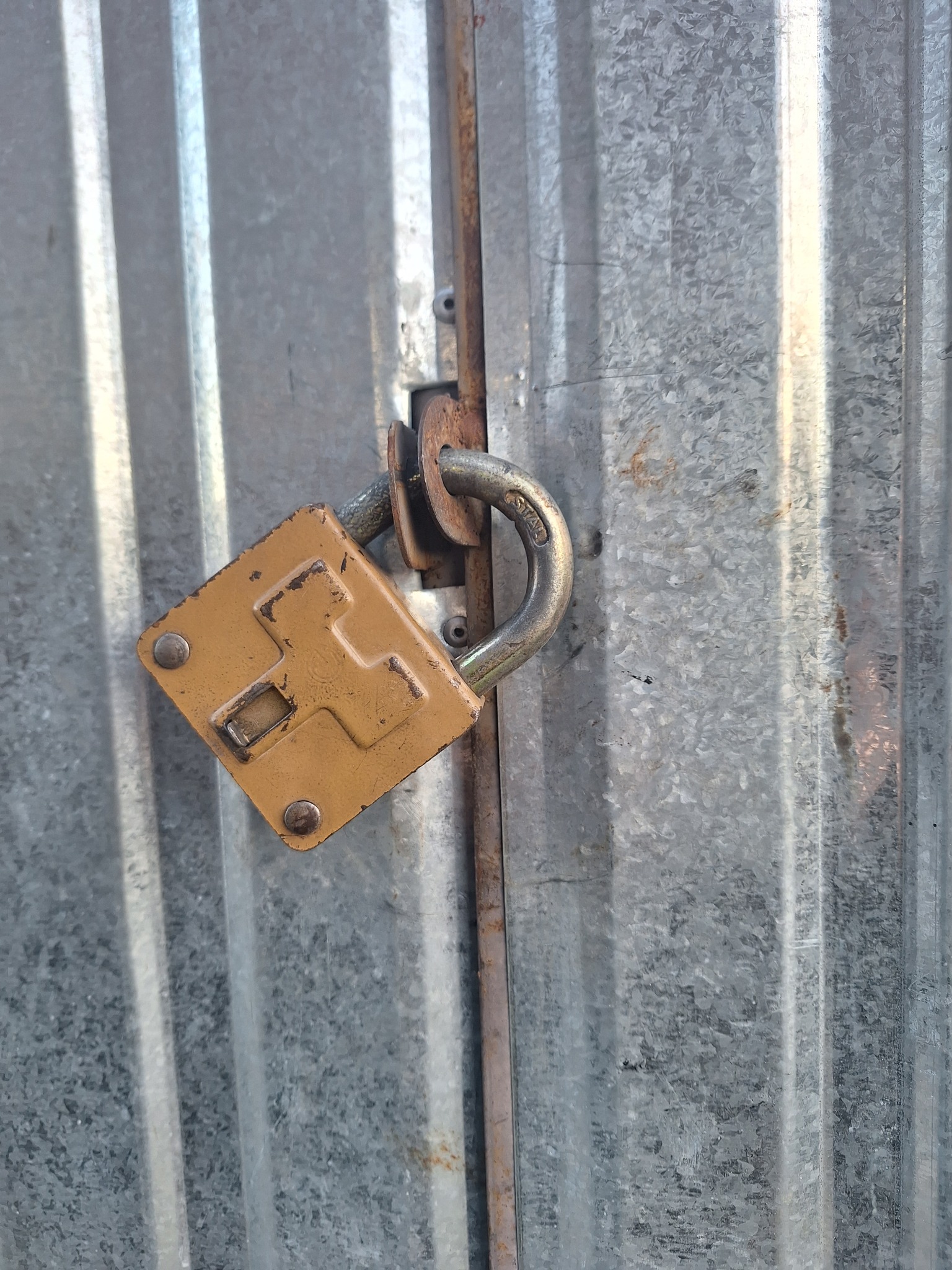 Zardzewiały, kwadratowy zamek na metalowych drzwiach z blachy falistej, zabezpieczający wejście.
