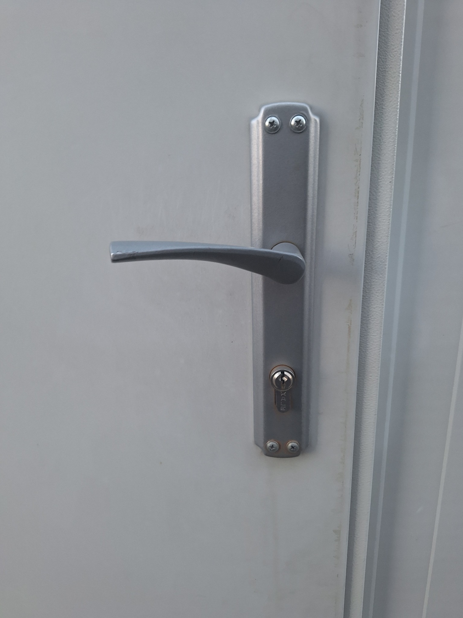 Szare drzwi metalowe z zamontowanym srebrnym zamkiem z klamką, widoczne ślady użytkowania na powierzchni drzwi.