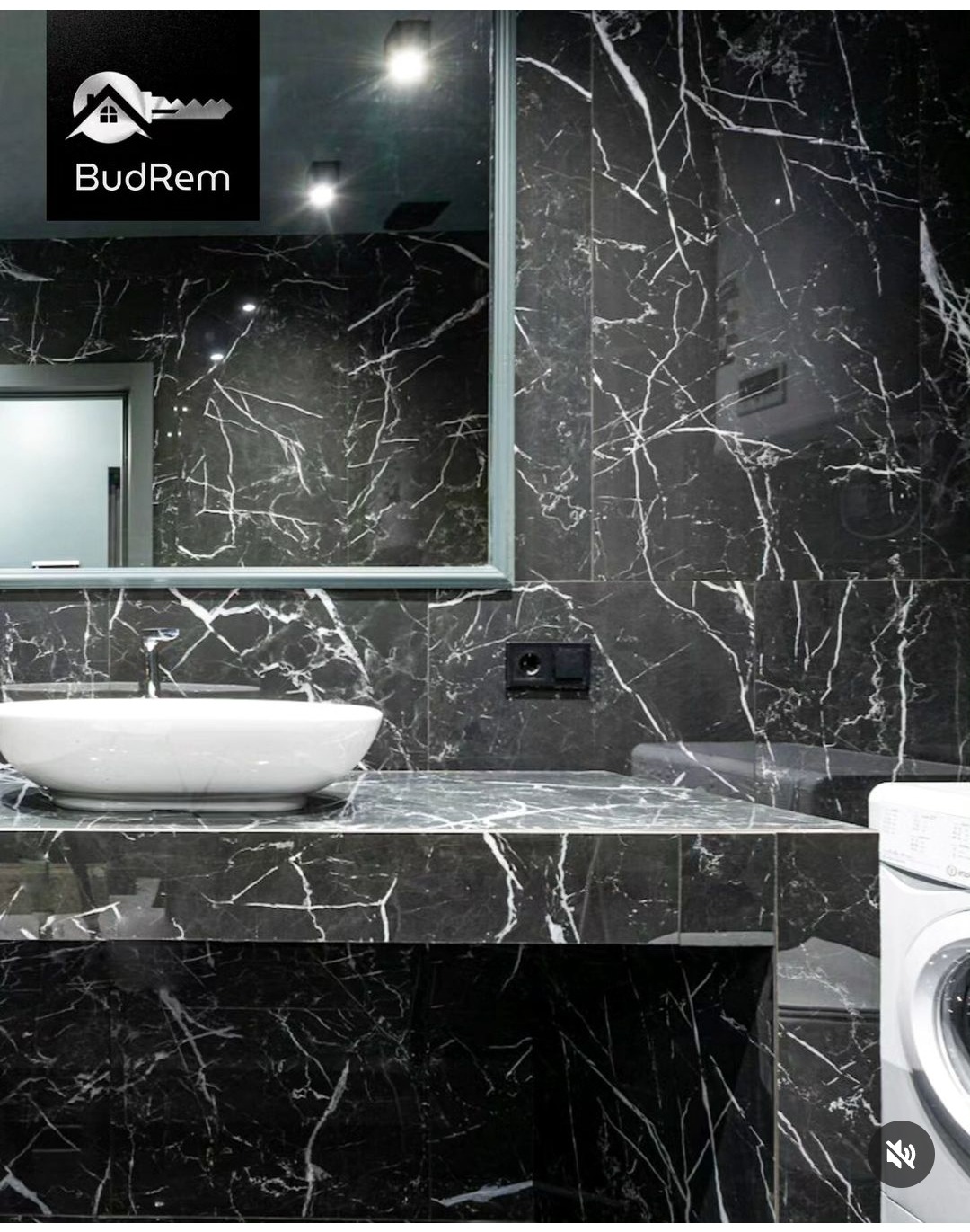 Elegancka łazienka z czarnymi marmurowymi płytkami z białymi żyłami, białą owalną umywalką nablatową, dużym lustrem i częściowo widoczną pralką.