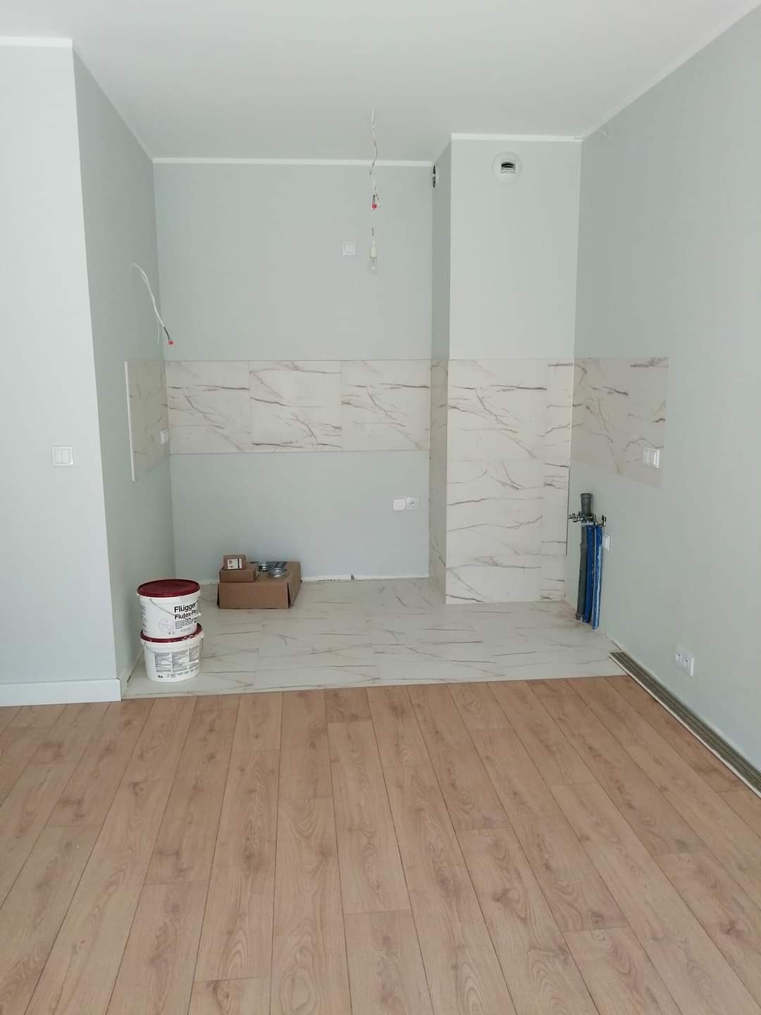 Wnętrze w trakcie wykańczania: położone panele podłogowe, częściowo ułożone płytki imitujące marmur na ścianie, puszki z farbą i karton na podłodze, widoczne instalacje elektryczne i hydrauliczne.