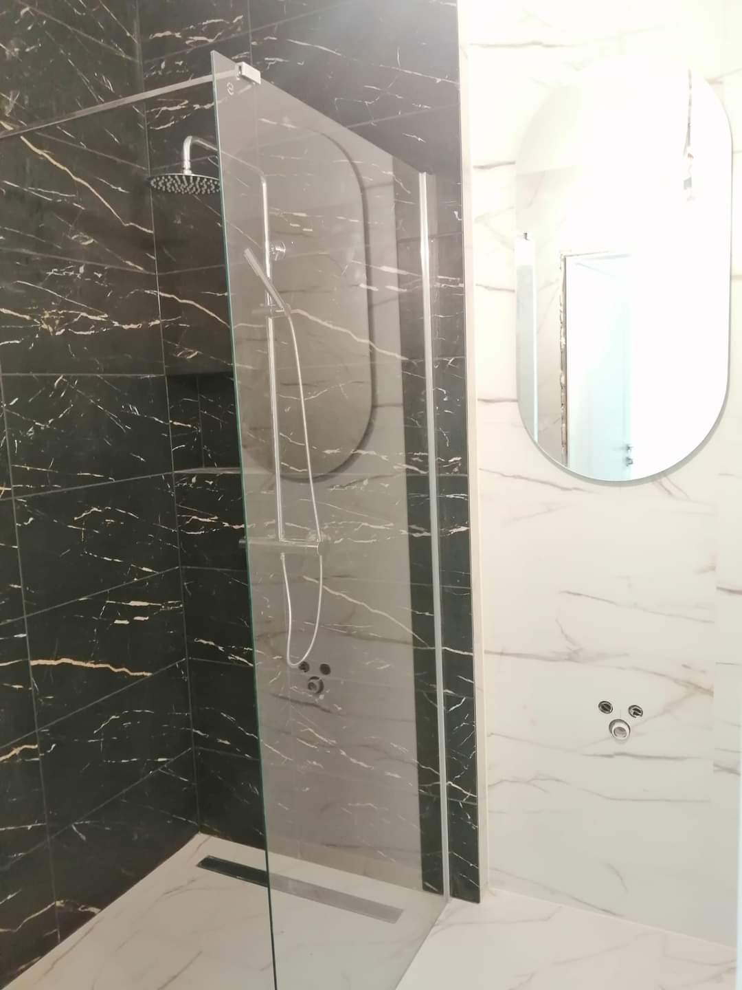 Nowoczesna kabina prysznicowa typu walk-in z czarnymi płytkami imitującymi marmur z złotymi żyłami, szklana ściana z chromowanymi profilami, owalne lustro na białej ścianie z marmurowym wzorem...